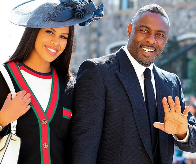 A smiling couple in formal attire at a public event, with the man waving and the woman wearing a large hat.