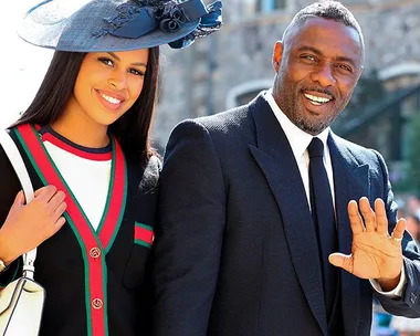 A smiling couple in formal attire at a public event, with the man waving and the woman wearing a large hat.