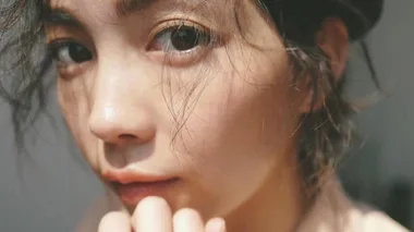 Close-up of a woman's face with natural makeup and tousled hair, looking directly at the camera in soft lighting.