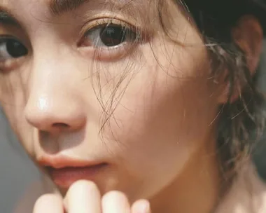 Close-up of a woman's face with natural makeup and tousled hair, looking directly at the camera in soft lighting.