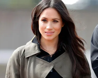 A woman with long dark hair, wearing a trench coat, posing outside with a neutral expression.