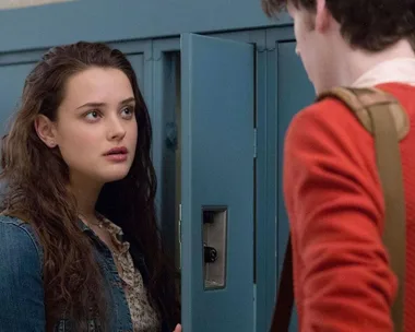 Teen girl stands by school locker, talking to a person with a back-facing view, in a scene from "13 Reasons Why."