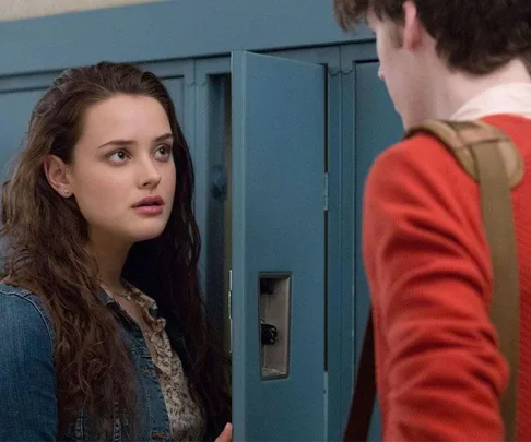 Teen girl stands by school locker, talking to a person with a back-facing view, in a scene from "13 Reasons Why."