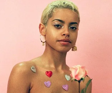 A person with short blonde hair, heart stickers on chest, holding a rose against a pink backdrop.