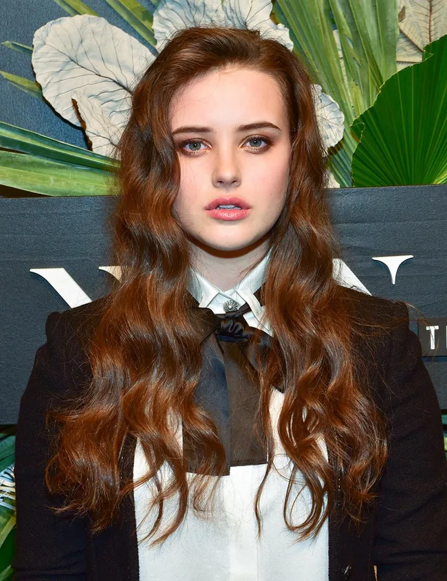 Young woman with long, wavy brown hair in formal attire, standing against a leafy background.