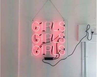 Neon sign reading "GIRLS GIRLS GIRLS" in pink, hanging on a wall with black power cord.