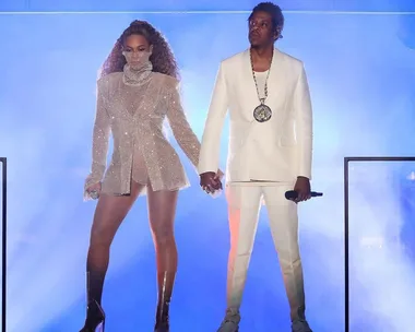 Beyoncé in a sparkling outfit and Jay-Z in a white suit hold hands on stage with a blue-lit background during a performance.