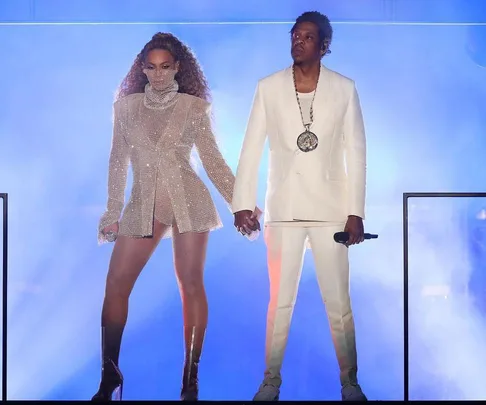 Beyoncé in a sparkling outfit and Jay-Z in a white suit hold hands on stage with a blue-lit background during a performance.