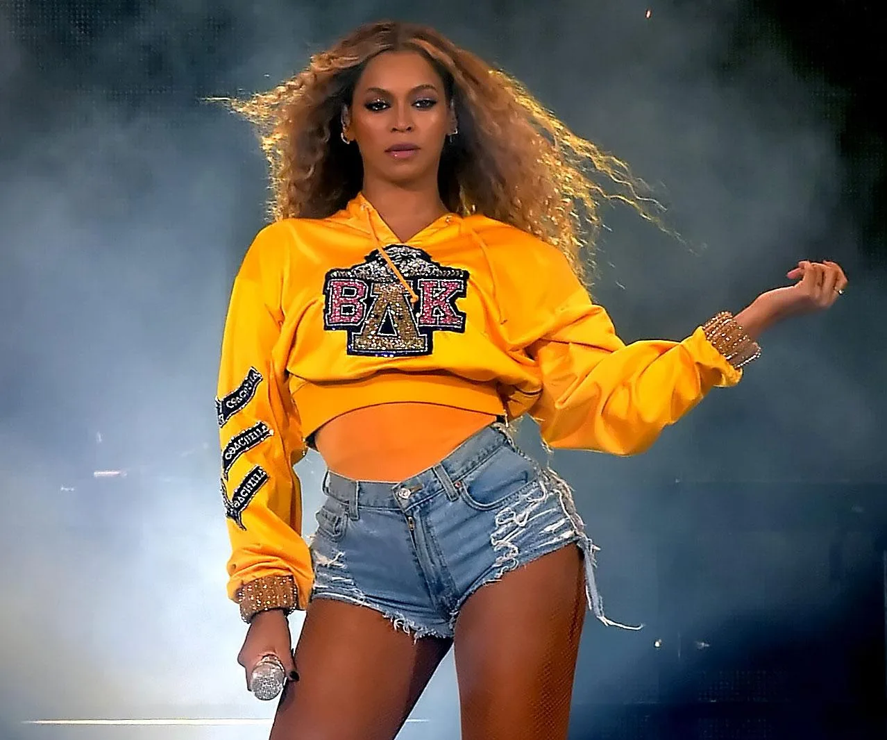 Beyoncé performs in a yellow sweatshirt and denim shorts, holding a microphone, with a smoky background.