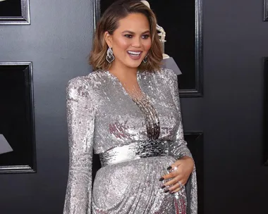 Chrissy Teigen in a silver dress, smiling and posing with one hand on her belly at an event.