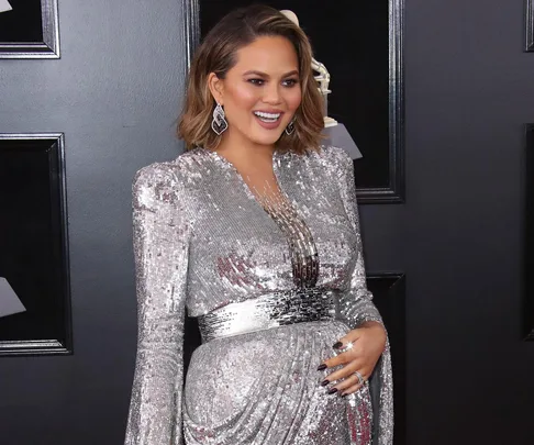 Chrissy Teigen in a silver dress, smiling and posing with one hand on her belly at an event.