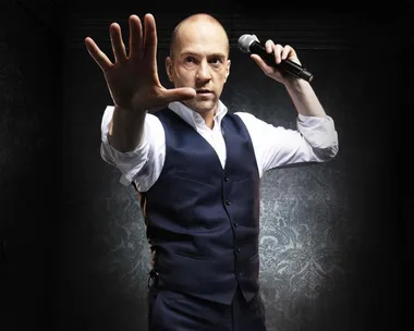 "Performer in a vest holding a microphone, reaching out with one hand in a dramatic pose."