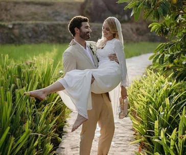 Man in beige suit carrying woman in wedding dress on a path surrounded by greenery.