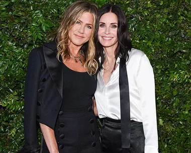 Two women in black and white outfits smile, standing in front of a leafy backdrop.