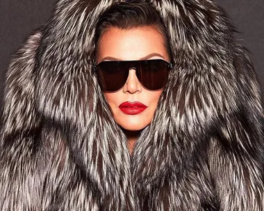 A woman wearing large black sunglasses and red lipstick, dressed in a luxurious fur coat, staring straight ahead.