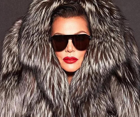 A woman wearing large black sunglasses and red lipstick, dressed in a luxurious fur coat, staring straight ahead.