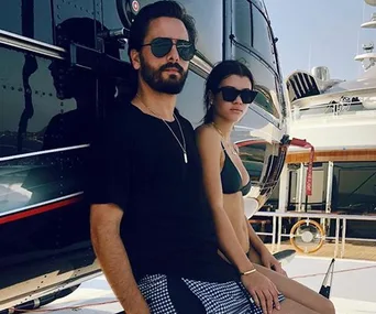 A man in a black shirt and sunglasses and a woman in a bikini and sunglasses stand near a helicopter on a yacht deck.