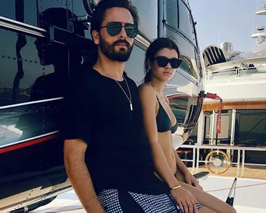 A man in a black shirt and sunglasses and a woman in a bikini and sunglasses stand near a helicopter on a yacht deck.