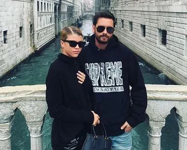 A couple stands on a bridge over a canal, both wearing dark sunglasses and cozy clothing.