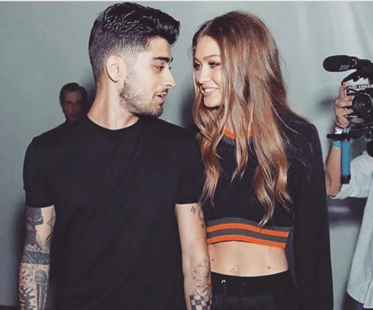 Gigi Hadid and Zayn Malik walking together, smiling at each other, with a cameraman capturing the moment.