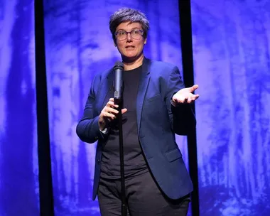 Hannah Gadsby performing on stage in "Nanette," wearing a blue blazer, glasses, and holding a microphone, with a blue background.