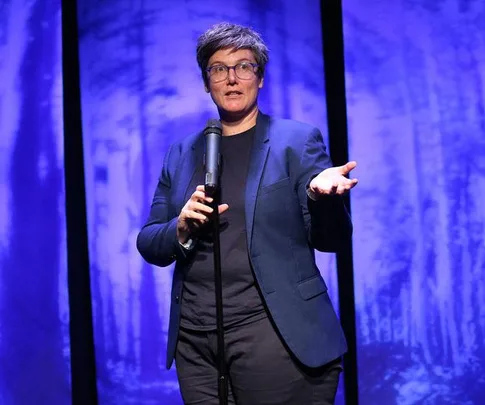 Hannah Gadsby performing on stage in "Nanette," wearing a blue blazer, glasses, and holding a microphone, with a blue background.