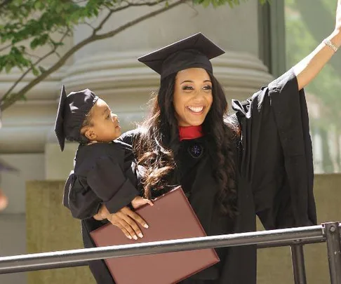 A woman in graduation gown holds a baby in matching outfit, both smiling and celebrating outdoors.