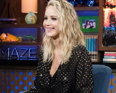 Jennifer Lawrence smiling while seated indoors, wearing a black patterned blouse with wavy blonde hair.