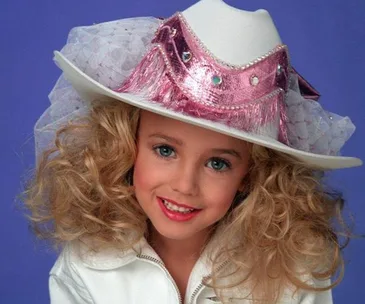 A blonde girl in a white cowgirl hat adorned with pink sequins and fringe, wearing a white outfit, smiling against a blue background.