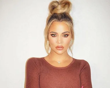 Woman with blonde hair in a top bun, wearing a brown top and a cross necklace, posing against a plain background.