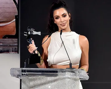 Kim Kardashian West holding CFDA Influencer Award, standing at a podium in a white outfit, speaking into microphones.
