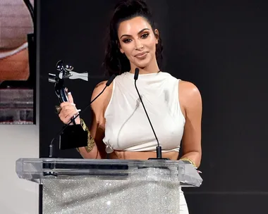 Kim Kardashian holding an award statue while standing behind a glittering podium, dressed in a white outfit.