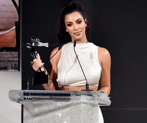 Kim Kardashian holding an award statue while standing behind a glittering podium, dressed in a white outfit.