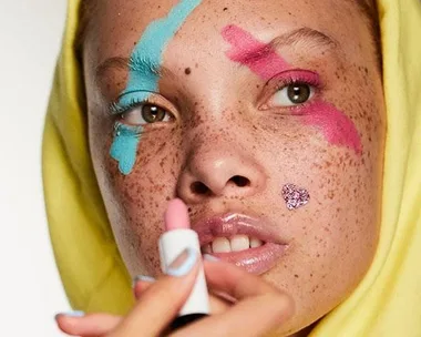 Close-up of a person with blue and pink makeup on their eyebrow and eye, wearing a yellow hoodie and holding a lipstick.