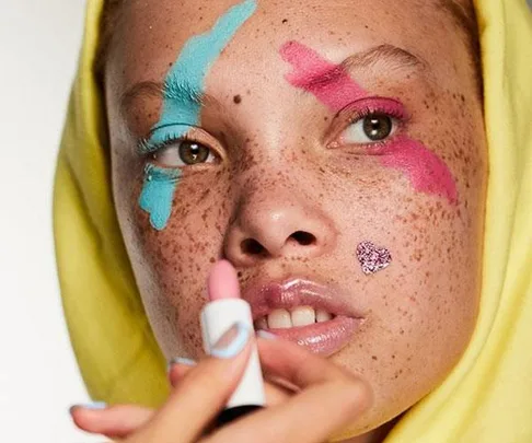 Close-up of a person with blue and pink makeup on their eyebrow and eye, wearing a yellow hoodie and holding a lipstick.