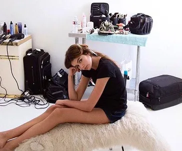 Young woman sitting on a fluffy stool in a makeup room with beauty products and suitcases around her.