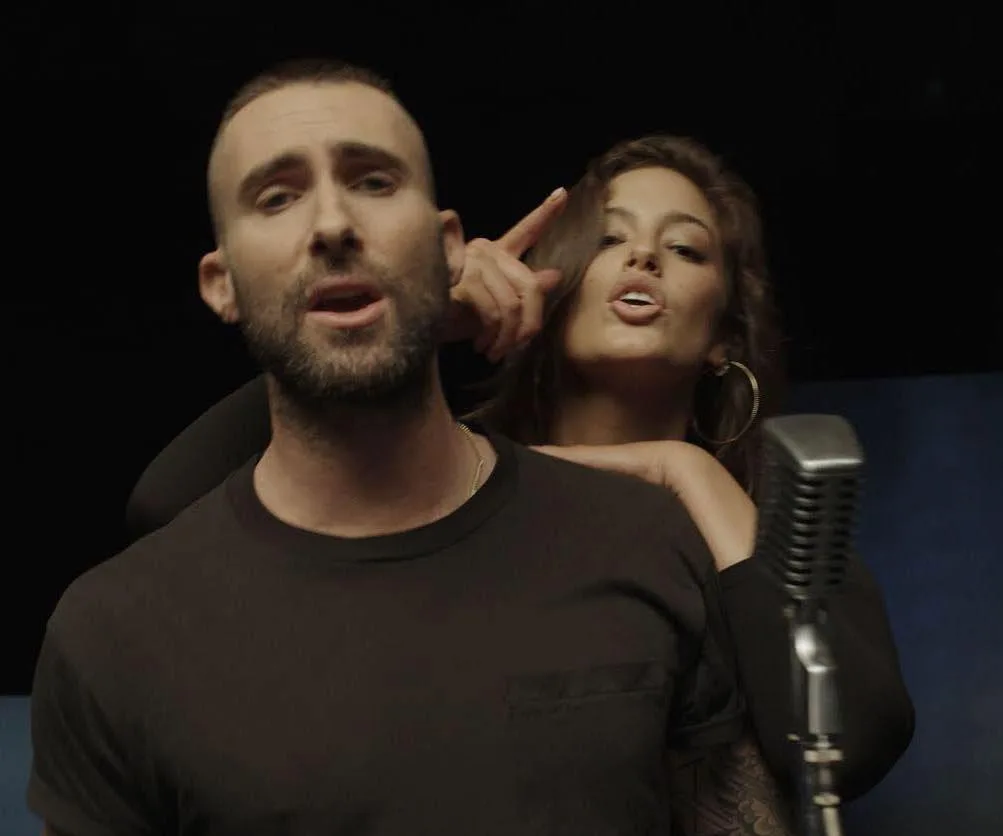 Singer with a beard sings at a microphone, accompanied by a woman gesturing playfully behind him.