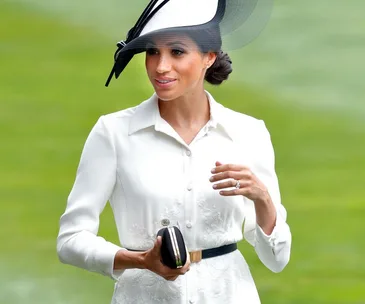 Woman in white dress with black belt and hat, holding a black clutch bag.