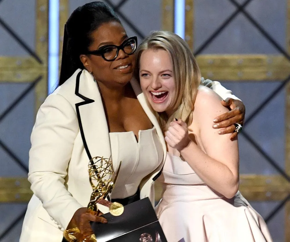 Oprah Winfrey hugs Elisabeth Moss, who is holding an Emmy award, both smiling on stage.