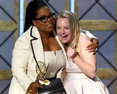 Oprah Winfrey hugs Elisabeth Moss, who is holding an Emmy award, both smiling on stage.