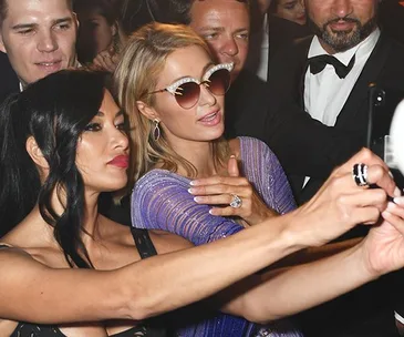 Paris Hilton taking a selfie with friends, showcasing her engagement ring, wearing a sparkling outfit and sunglasses at an event.