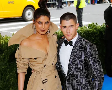 Nick Jonas and Priyanka Chopra at an event; she wears a beige trench coat, he wears a patterned suit with a bow tie.