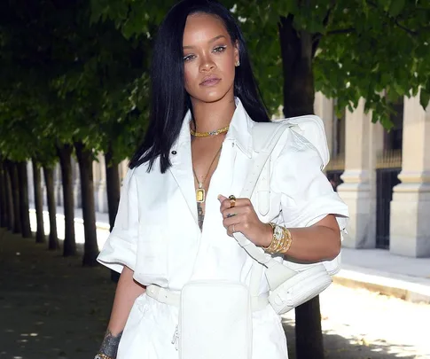 Woman in white outfit with gold jewelry, carrying white bag, standing outdoors with trees and buildings in the background.