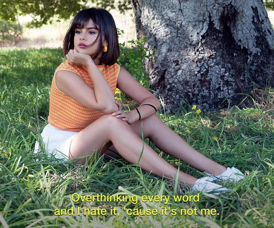 A young woman with short dark hair sits on grass in front of a tree, looking pensive. Subtitled text: "Overthinking every word and I hate it, 'cause it's not me."