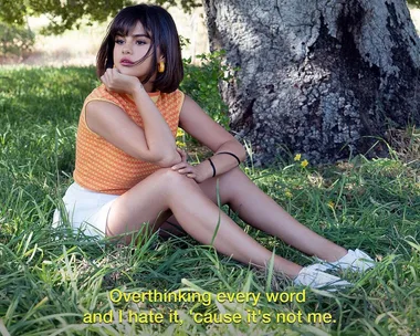 A young woman with short dark hair sits on grass in front of a tree, looking pensive. Subtitled text: "Overthinking every word and I hate it, 'cause it's not me."