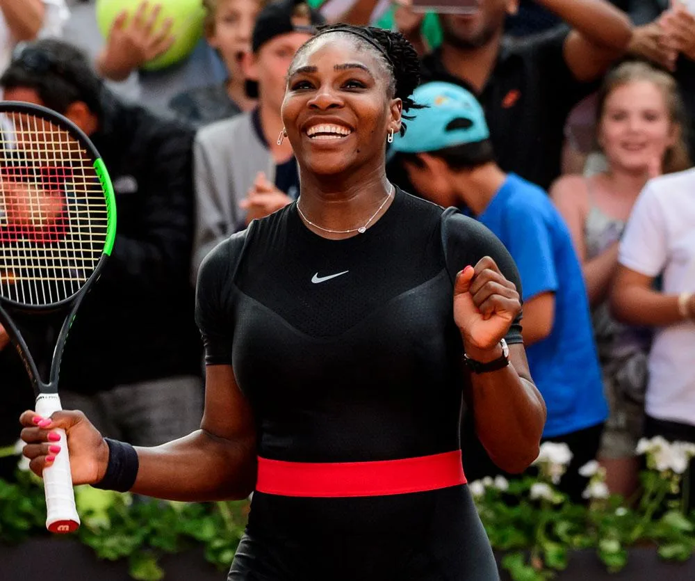 A tennis player smiling and holding a racket, wearing a black outfit with a red belt, surrounded by cheering crowd.