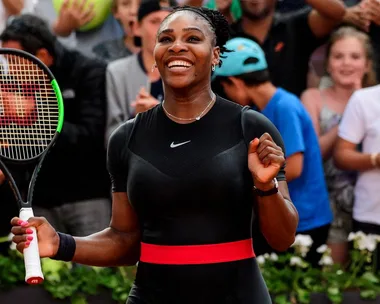 A tennis player smiling and holding a racket, wearing a black outfit with a red belt, surrounded by cheering crowd.