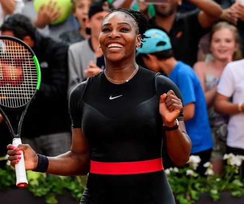 A tennis player smiling and holding a racket, wearing a black outfit with a red belt, surrounded by cheering crowd.