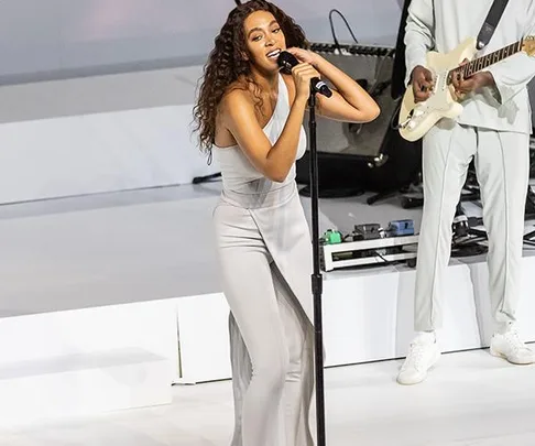 Solange performing on stage in a light gray outfit, singing into a microphone with a guitarist in the background.