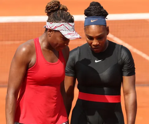 Two female tennis players in conversation on a clay court, one in a red tank top and the other in a black outfit.